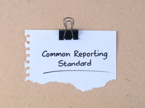 White torn paper note with handwritten text common reporting standard secured by a black binder clip on brown cardboard texture surface, financial and tax compliance concept