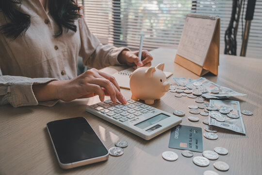 Woman calculating monthly expenses on calculator beside credit card, piggy bank, cash, and calendar for financial debt management