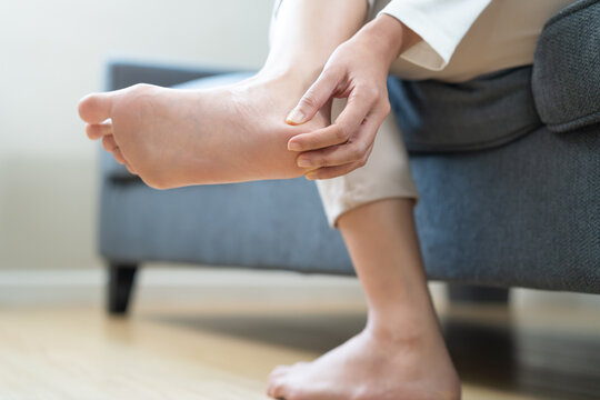 Foot pain concept, close up hand of young woman rubbing, massaging sore feet area of heel, girl suffering on sofa, couch at home. Discomfort painful feet ache from walking for long. Physical injury.