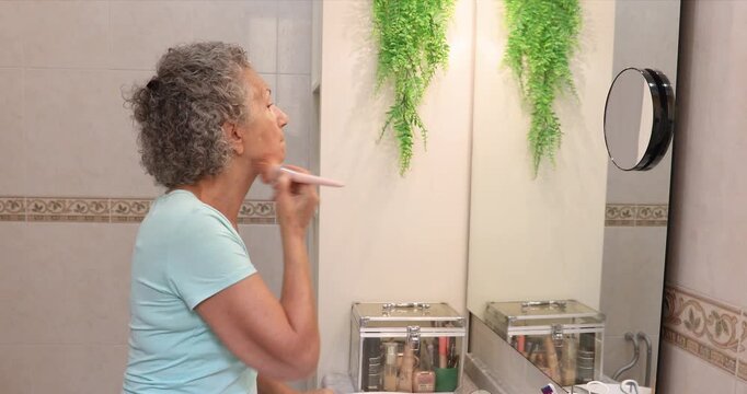Mature woman applying foundation with a makeup brush while looking into a bathroom mirror during her daily beauty routine.