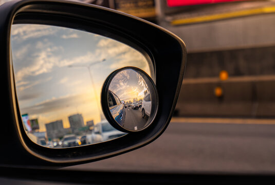 Car side mirror reflecting traffic on urban road at sunset, with convex blind spot mirror showing nearby vehicles for safer driving, road awareness and transportation safety. Accident prevention.