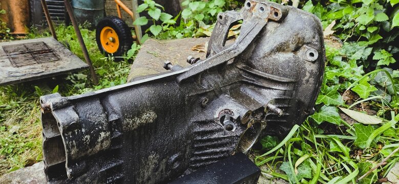 A worn mechanical housing lies in the grass, seemingly abandoned among leaves and garden tools. Bolts, corroded surfaces, and industrial shapes stand in sharp contrast to the softness of the natural 