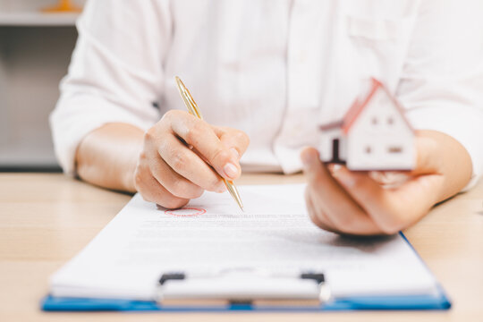 Person holding a miniature house and pointing to financial documents with a pen symbolizes mortgage consultation, home loan review, property purchase, contract terms, financing,