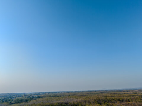 Vast clear blue sky over a rural landscape with a small village, green fields, and dry forest horizon.