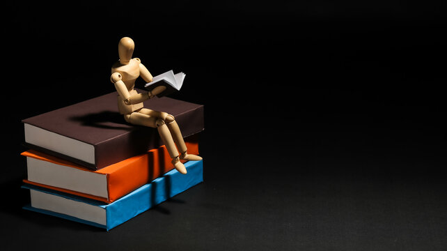 Wooden mannequin sitting on a stack of big books while reading a small book, symbolizing education, knowledge, self improvement, or lifelong learning.
