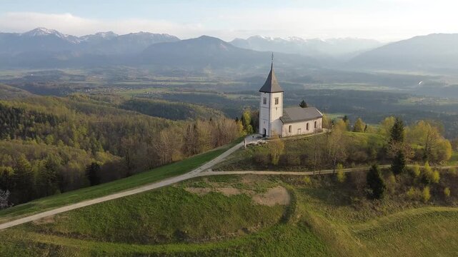 Aerial view of Sts. Primus and Felician Church in Jamnik, Slovenia, standing on a green hill surrounded by spring forest, with a scenic valley and snow-capped Julian Alps in the background.

