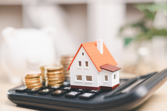 Miniature house and coin stacks on a calculator symbolize mortgage calculation, home affordability, property costs, loan payments, interest rates, budgeting, and real estate finance planning.
