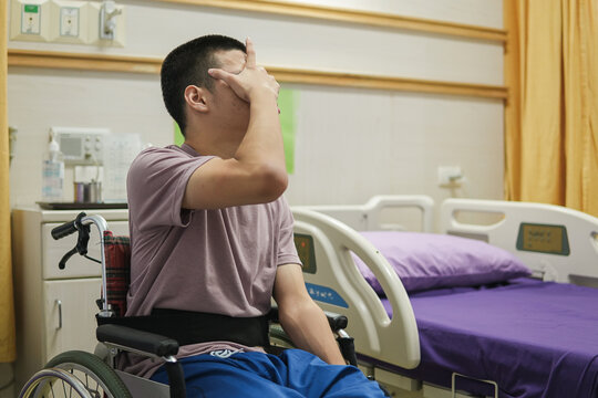 Young man in a wheelchair sitting alone in a hospital room, covering his face with his hand, expressing sadness, stress and emotional distress during illness, treatment and recovery