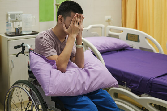 Young disabled man in a wheelchair sitting alone in a hospital room, covering his face with his hands, expressing sadness, anxiety and emotional distress during illness and treatment
