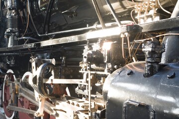 Close-up of vintage steam locomotive undercarriage revealing black machinery and red wheels. Copper wiring gleams in sunlight, highlighting industrial engineering for heritage railway themes. © Grenar