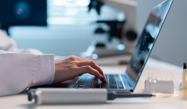 Scientist works on a laptop in a modern laboratory with a micropipette, sample rack, and microscope nearby, illustrating research documentation, data analysis, biotechnology, and digital science.
