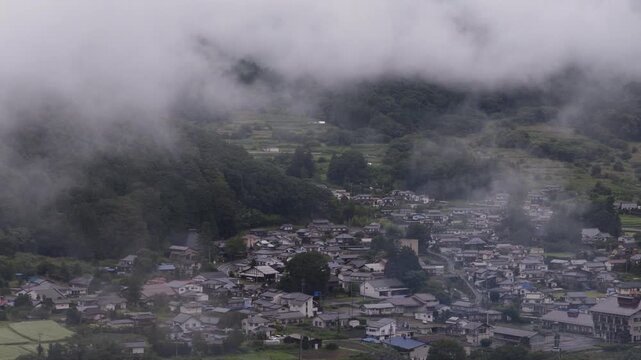 Fog and clouds move over small town at foot of green mountains in Nagano, Japan