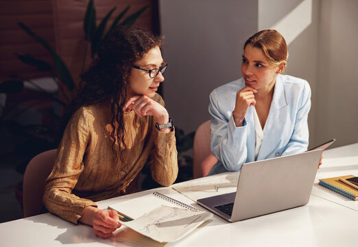 Two caucasian women rest their chins on their hands, facing each other across a sunlit white desk, where printed line charts, a notebook and a laptop lie between them.