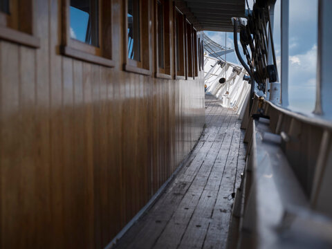 Narrow side deck running alongside a varnished wooden deckhouse with portlights, coiled lines and blocks hanging along the rail.