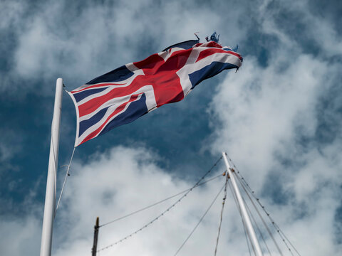 The Union Jack lifts diagonally from the mast of a sailing ship, its flying edge worn thin against blue sky and white clouds.