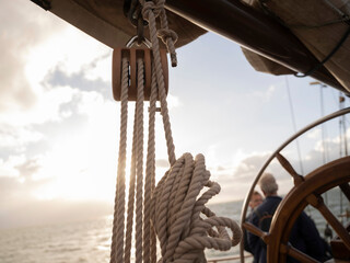 A wooden triple block with rope hangs below the boom of a sailing ship while two passengers talk near the wheel in warm light. © Victor