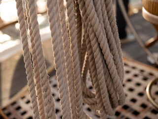 A bundle of twisted natural ropes hangs above a wooden deck grating on a sailing ship, warm light raking across the fibres. © Victor