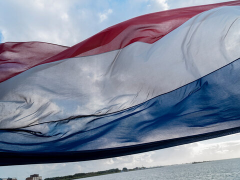 The Dutch flag streams in the wind above open water, sunlight shining through the fabric with a low coastline behind.