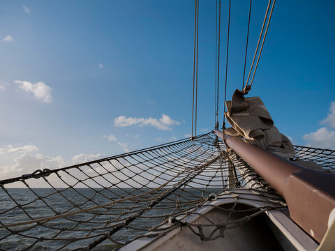 Looking out along the bowsprit of a sailing ship with its net and furled jib towards a calm sea and a wide blue sky.