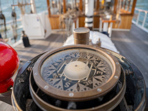 A brass compass in its binnacle with a red sphere, looking forward along the wooden deck of a sailing ship towards the masts.
