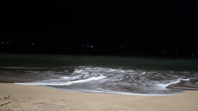 Night shot of a sandy beach as sea waves roll in. The surging wave stirs up sand from the bottom, while colliding undercurrents and incoming waves crash into each other, creating thick white foam.