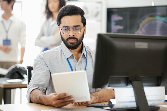 Business professional using a tablet while working at a computer workstation, with colleagues and digital information in the background.