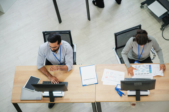 Top view of business professionals working at computer workstations and reviewing charts, reports, and digital information in a modern office.