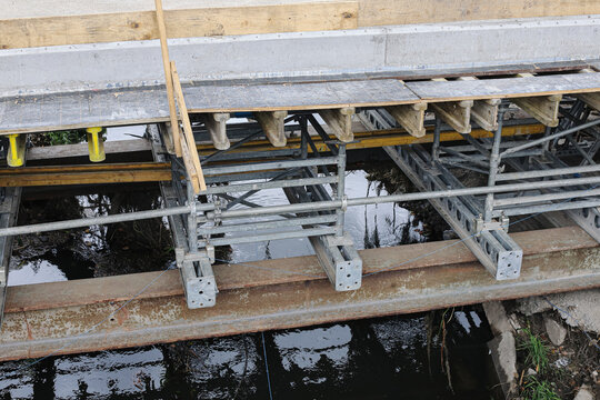 Bridge construction above water. Metal scaffolding supports concrete. Wooden formwork covers section. Steel beams below platform. Temporary supports over water. Construction background with framework.