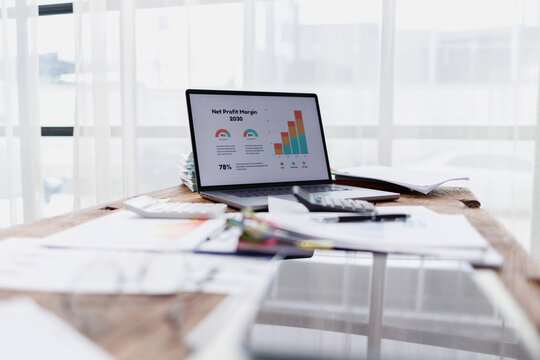 Laptop displaying net profit margin 2030 financial data charts on its screen, surrounded by calculators and documents on a wooden desk, symbolizing business analysis and growth