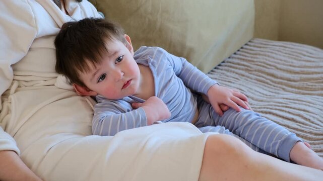 Toddler boy resting on lap of mother in cozy bedroom. Caring woman holding baby son on couch at home. Peaceful morning scene representing childhood, maternity, love and family bonding.