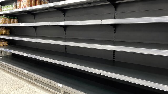 Empty black metal supermarket shelves with a few packaged goods on the top left, long retail aisle and tiled floor under fluorescent lighting