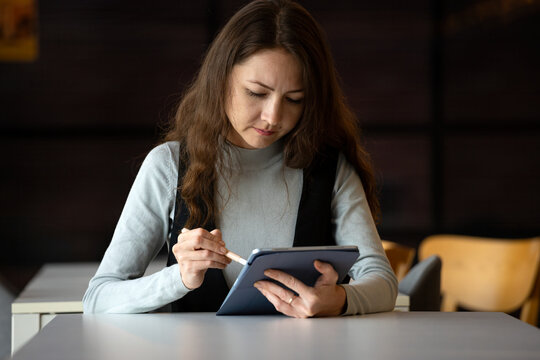 Focused woman using tablet and stylus at desk in modern workspace, showing digital work, productivity, technology, concentration, learning and professional creative activity
