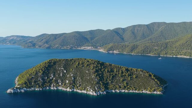 Aerial view of a lush, green, forested island surrounded by deep blue sea, with the mountainous coastline of Skopelos island and the village of Neo Klima visible in the background, Greece