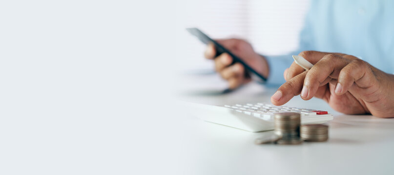 Businessman using calculator and mobile phone with coin stacks on white desk with copy space, concept of financial calculation, personal finance, budget planning, and accounting.