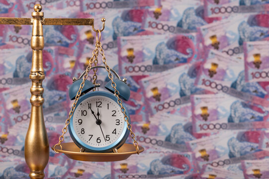 An alarm clock on the pan of golden scales against a background of 10,000 Kazakhstani tenge banknotes.