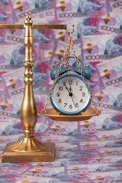 An alarm clock on the pan of golden scales against a background of 10,000 Kazakhstani tenge banknotes.