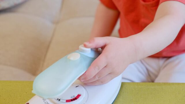 Toddler playing with a small white plastic iron. Child hands touching household appliance toy. Children learning through pretend play. Development of motor skills and imagination.