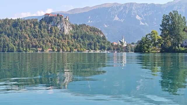 Scenic view across Lake Bled toward Bled Castle and St. Martin's Parish Church, with forested hills, mountains and colorful autumn foliage reflected in the water.