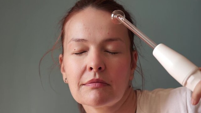 woman using darsonval electric device on cheek gazing away in bright studio closeup, concept of skincare routine, cosmetology, aesthetic medicine. High quality photo