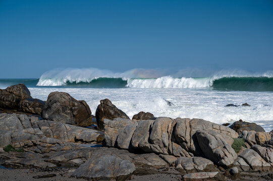 Powerful ocean waves break in succession along a rugged rocky coastline, sending white spray into the air beneath a clear blue sky. The rolling surf and emerald-green wave faces create a dramatic seas