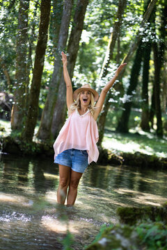 Happy woman wading in a sunlit river, arms raised and splashing water, enjoying carefree summer nature and joyful freedom