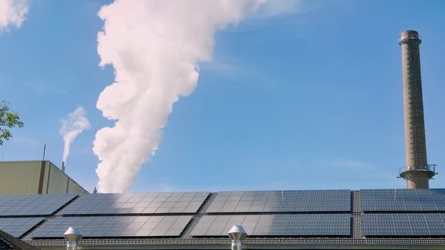 Industrial building with rooftop solar panels and steam rising from a tall chimney against a clear blue sky, highlighting renewable energy and industrial infrastructure