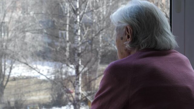 Elderly woman with grey hair gazing out of window at winter landscape. Old female reflecting on life with sense of loneliness and solitude in nursing home or apartment