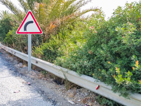 Triangular road sign with a black curved arrow symbol stands firmly beside a metallic guardrail on a sunny day near lush green shrubs and palm trees.