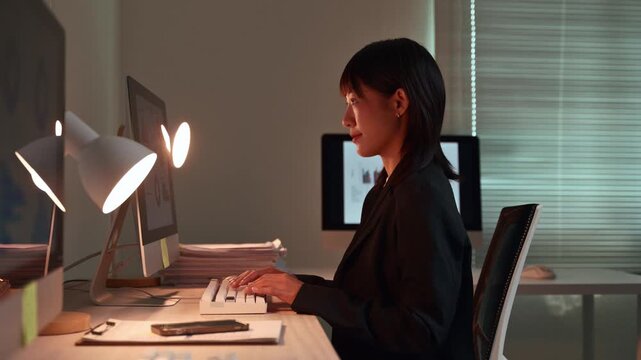Focused young asian businesswoman working late at night in a dark office, typing on a keyboard and looking at a computer monitor with charts and data to meet a project deadline