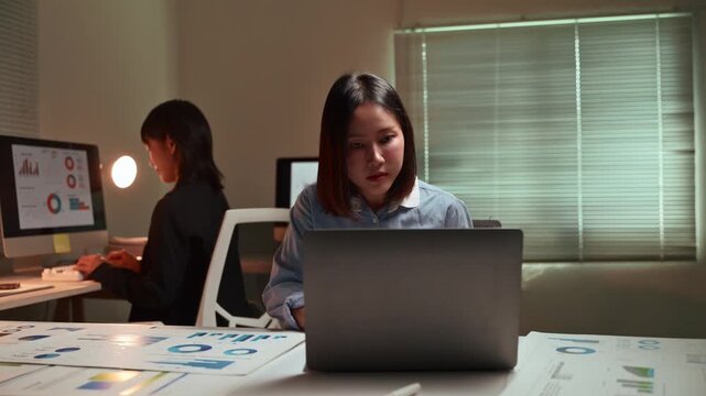 Young Asian woman working late at night on a laptop computer, with a colleague in the background analyzing charts and graphs on her screen, demonstrating dedication and overtime commitment