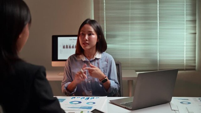 Two young Asian businesswomen discussing a financial project and analyzing market data charts on paperwork while working overtime together in a modern office at night