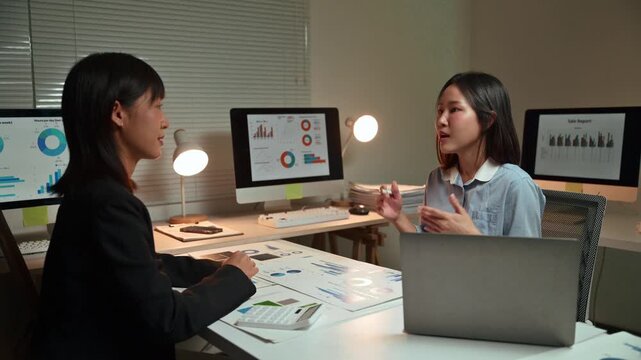 Two Asian businesswomen discussing a new project, brainstorming ideas, and reviewing financial data on documents and computer screens while working together late in the modern office