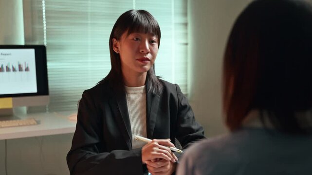 Serious asian woman in a suit presenting a business proposal to a colleague during a late-night meeting in a modern office, gesturing and explaining data from a sales report on her desk