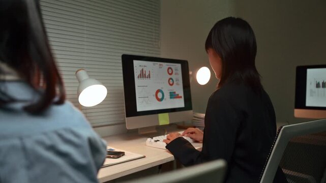 Young Asian businesswoman using a desktop computer to analyze financial data charts and graphs, pointing at the screen while working overtime late at night in a modern office with her colleague